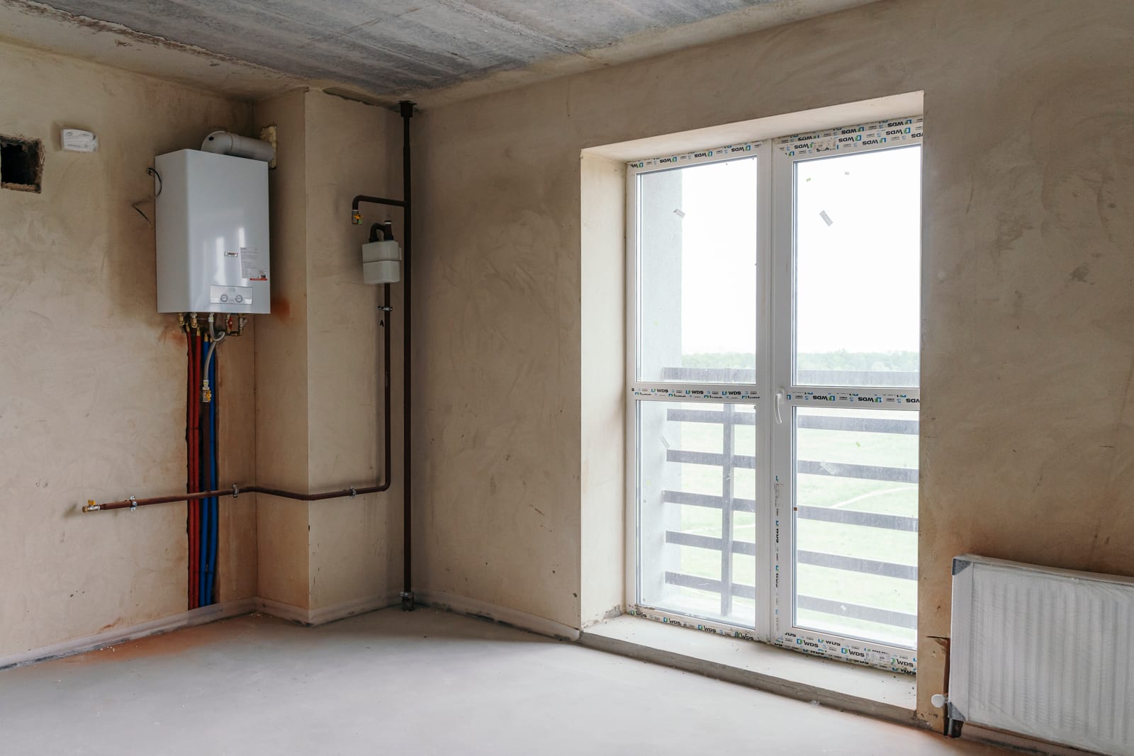 Una habitación entregada sin acabados: solera, enlucido, un radiador, la caldera y una ventana a campo abierto