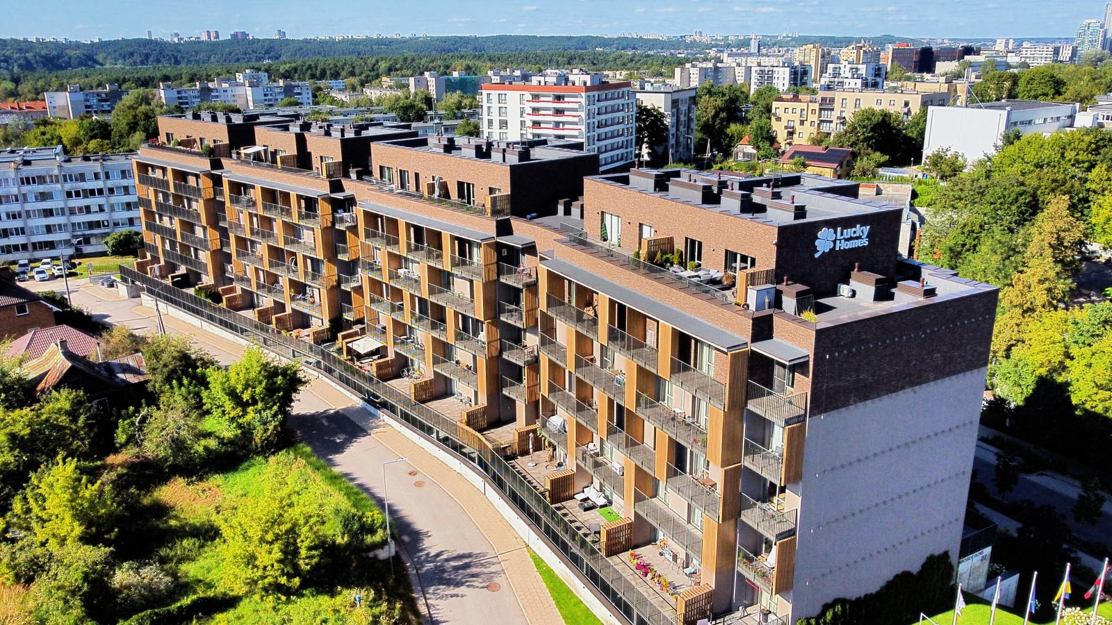 Lucky Homes by SAVO Vilniuje iš oro: apšviestas pavadinimas ant galinės sienos, apželdintos stogo terasos ir Vilniaus panorama horizonte