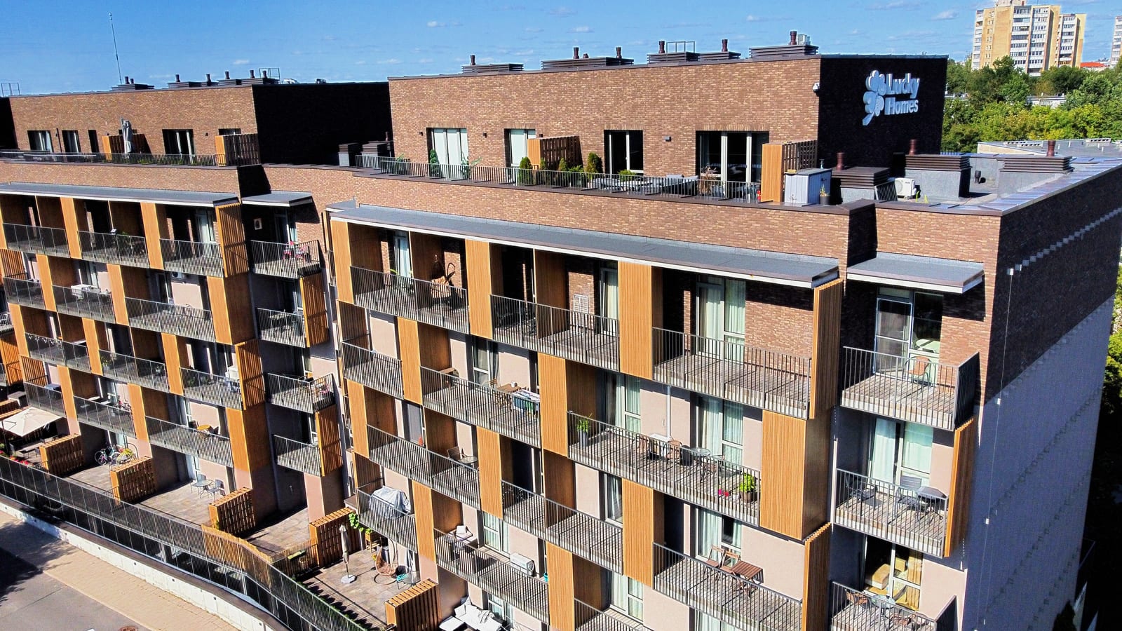 Luftaufnahme entlang des Gebäudes mit dem beleuchteten Schriftzug Lucky Homes an der dunklen Giebelwand, darunter die Balkone zwischen Klinkerpfeilern und Paneelen aus Holzlamellen.